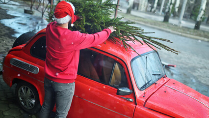A young man in a red sweater and a Santa hat is tying a Christmas tree to the roof of a red retro car.