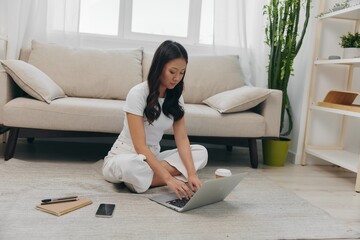 Young Asian female student looking at laptop screen and studying for college exams sitting on the floor at home with notebooks to write, lifestyle freelance work