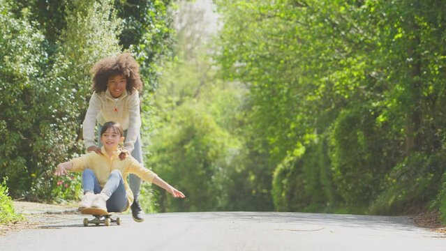 Children having fun outdoors with boy pushing girl on skateboard along country road