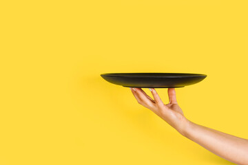 Mano de mujer sosteniendo un plato negro sobre un fondo amarillo brillante liso y aislado. Vista de frente y de cerca. Copy space