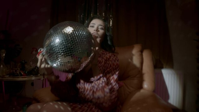 Brunette girl sitting on a brown sofa holding a disco ball 