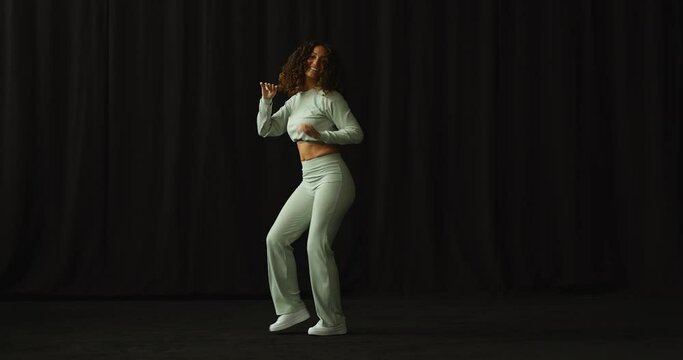 An beautiful young african american woman with big curly hair wearing a green dance outfit dancing bachata in a all black dance studio. 