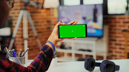 Female freelancer holding smartphone with horizontal greenscreen in living room, using blank copy...
