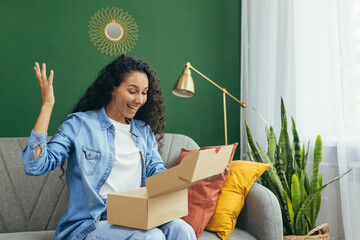 Young beautiful curly haired hispanic woman received package from online store, happy and satisfied with delivery sitting on sofa and happy in living room with green wall.