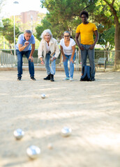 A group of multiracial mixed-age adult people lined up in a row at play of petanque game outdoors...