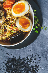 Sopa ramen, porchetta y huevo en tazón blanco, sobre un plato negro y a un lado semillas de sésamo en un fondo gris, vista superior