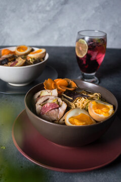 Platos de Sopa ramen, porchetta y huevo en taz&oacute;n blanco, sobre un plato negro y a un lado vaso de jugo de Jamaica