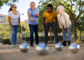 Middle aged mature white man throwing a boule ball in petanque game in the park with other people standing beside and observing him