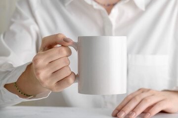 Fototapeta na wymiar Female hand holding white mug mockup with blank copy space for your advertising text message or promotional content. Girl in white shirt holding white porcelain coffee mug mock up