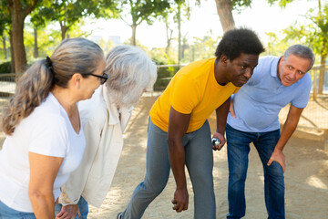 Middle aged mature black man throwing a boule ball in petanque game outside in park with other people standing beside