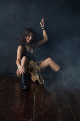 A brunette girl dressed in black shorts and a T-shirt, shod in boots, sits on the floor near a dark blue wall, dances and moves her arms. Creative state, mood to listen to music. Gothic brutal.