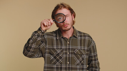 Investigator researcher scientist man holding magnifying glass near face, looking into camera with big zoomed funny eyes, searching, analysing. Young adult guy isolated on beige studio background