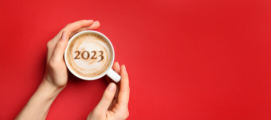 Female hands with cup of hot coffee and figure 2023 on red background with space for text