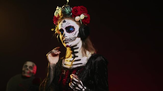 Lady of death listening to music on headphones, wearing skull make up and santa muerte costume on day of the dead. Enjoying fun song on audio headset, celebrating mexican tradition. Handheld shot.