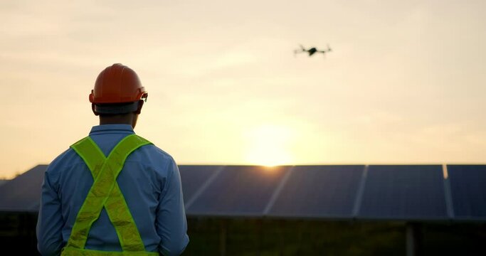 Backlit scene, Asian Young Inspector Engineer man wearing protection helmet control drone flying over row of solar panel while checking operation in solar station in evening