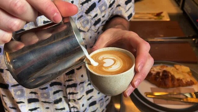 Professional barista hands gently pouring the frothed milk into a cup of coffee shot, breaking open the crema and making beautiful latte art.