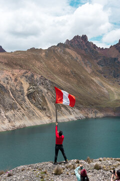 Un hombre alzando la bandera peruana en la monta&ntilde;a, a la orilla de una laguna, y dos mujeres tom&aacute;ndole fotos con el m&oacute;vil, y el cielo nublado, en los andes de PER&Uacute; Sudam&eacute;rica	