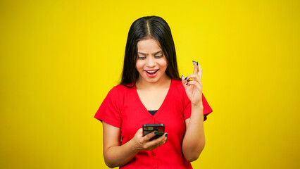 Portrait of a smiling young girl holding phone over yellow background, holding fingers crossed for good luck