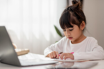 Fototapeta premium Asian schoolgirl doing her homework with laptop at home. Children use gadgets to study. Education and distance learning for kids. Homeschooling during quarantine. Stay at home