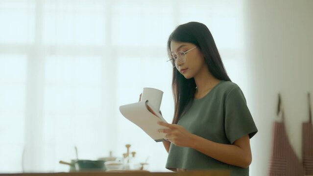 Happy young asian woman wearing glasses in kitchen at home. Female concentrate reading note cooking