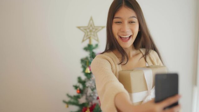 Young asian woman having fun taking light cheerful selfie and video call talking with her friend. Female showing gift box while on video conversation with friend. Happy new year, Thanksgiving