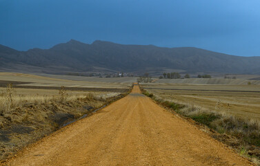 way forward in a flat landscape a smooth rustic road of yellowish earth advances straight towards the mountains on a sad day, leaden gray, on the sides empty arid deserted fields without trees, only w