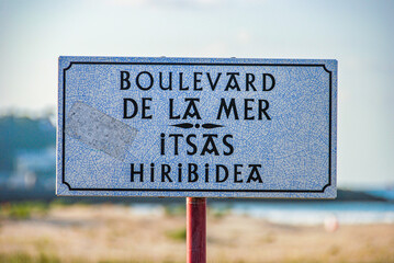 Name sign of a street, Avenida de la Mar, in French and Basque. Embossed white poster, with an antique, classic, Belle époque look. The card is in focus, the background is blurred. Boulevard de la mer