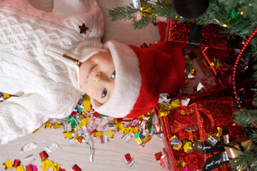 Happy smiling boy lying on floor in christmas santa hat near christmas tree with gifts. Caucasian preschooler waiting for new year