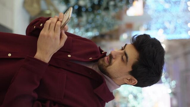 Vertical portrait of smiling man with beard typing message using phone looking on screen standing in hall of celebrate shopping mall in Christmas eve, on background of beautiful xmas decoration.