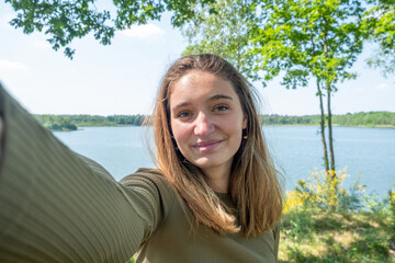 Beautiful young brunette Caucasian woman taking a selfie with smartphone outdoors on a forest lake in spring. High quality photo - obrazy, fototapety, plakaty