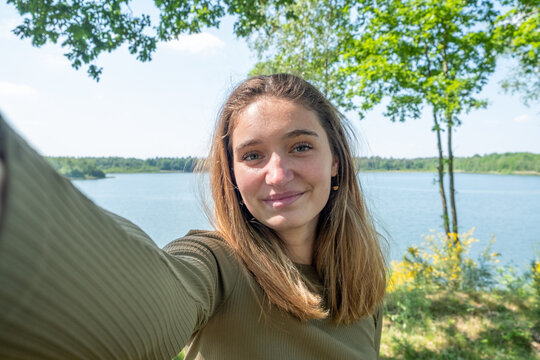Beautiful young brunette Caucasian woman taking a selfie with smartphone outdoors on a forest lake in spring. High quality photo