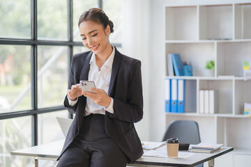 Young beautiful asian woman using smartphone and working with laptop while sitting at office desk, working from home concept.