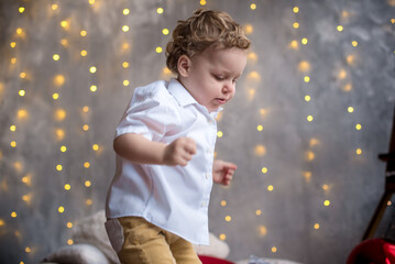 A little one and a half year old boy in pajamas sits under the Christmas tree and plays with a box with gifts. bokeh in the background from the New Year's garland