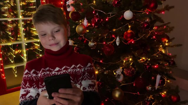 A cute smiling boy looks at the phone in the evning in front of the Christmas tree. leisure, technology and holidays concept