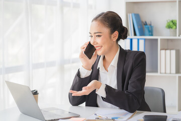 Young beautiful asian woman using smartphone and working with laptop while sitting at office desk, working from home concept.