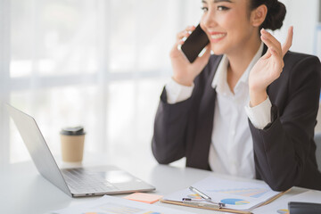 Young beautiful asian woman using smartphone and working with laptop while sitting at office desk, working from home concept.