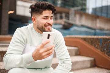 happy hispanic young man in sportswear using cellphone in urban scenario, big closeup view