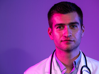 Coronavirus covid-19 danger alert Portrait of hero in white coat. Cheerful smiling young doctor with stethoscope in medical hospital standing against blue and pink background.