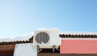 Snow covered invertor Air conditioner outdoor unit closeup on roof top. A snow-covered roof of a wooden house against blue sky background