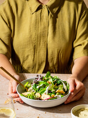 Woman in a green jumpsuit holding a plate with fresh salad, crackers, and hummus dressing on a table in front of her with hard shadows and minimal setting