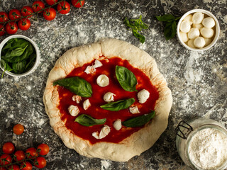 Raw dough with tomato sauce, mozzarella cheese balls, and fresh basil leaves, uncooked pizza Margarita top view on a blue marble background with flour in corner and ingredients