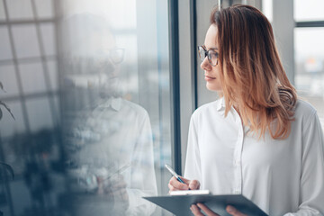 Young woman holding folder of documents and standing at the panoramic window of a modern office.