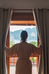 Vertical image of an unrecognizable woman with robe on her back drawing the curtains of her room...