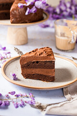 Slice of cake standing on a plate, chocolate brownie cake, with purple flowers around, and cup of coffee