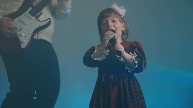 Little girl on stage in vintage dress, she sings into microphone and dances, her father plays an electric guitar. Color music is shining and smoke is billowing. Family time, creativity and hobbies
