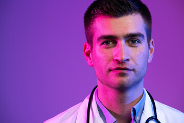 Obraz premium Coronavirus covid-19 danger alert Portrait of hero in white coat. Cheerful smiling young doctor with stethoscope in medical hospital standing against blue and pink background.