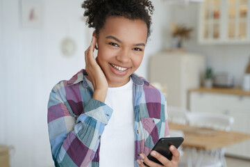 Smiling mixed race girl listens to music in earphone holding smartphone, using mobile apps at home