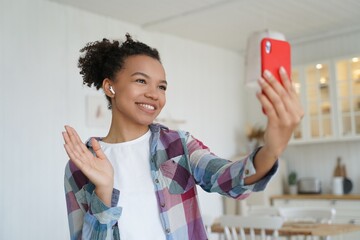 Smiling mixed race girl makes greeting hello gesture, talking by video call, holding phone at home