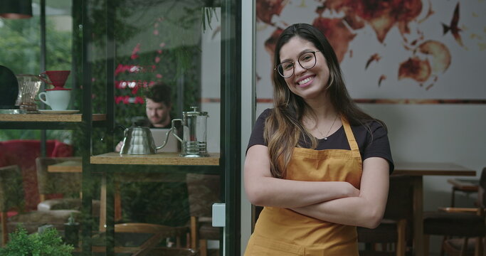 Happy female staff crossing arms smiling at camera in front of coffee shop store wearing yellow apron. A hispanic latin adult girl barista employee of small business restaurant place