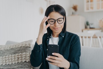 Modern businesswoman uses mobile apps on smartphone straightening glasses, sitting on sofa at home